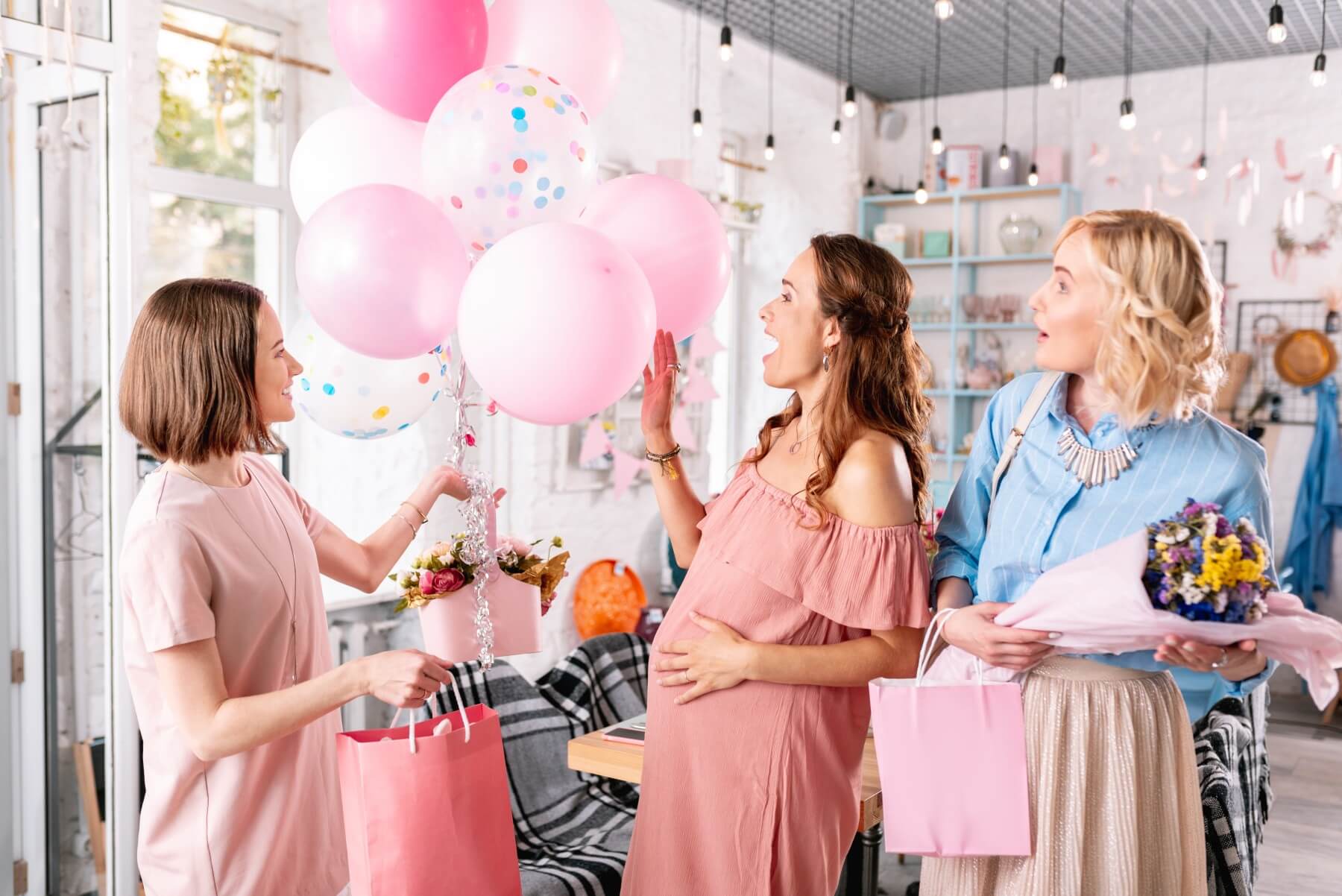 Babyshower feiring med gravide kvinner, rosa ballonger, gaver og pyntet festlokale.
