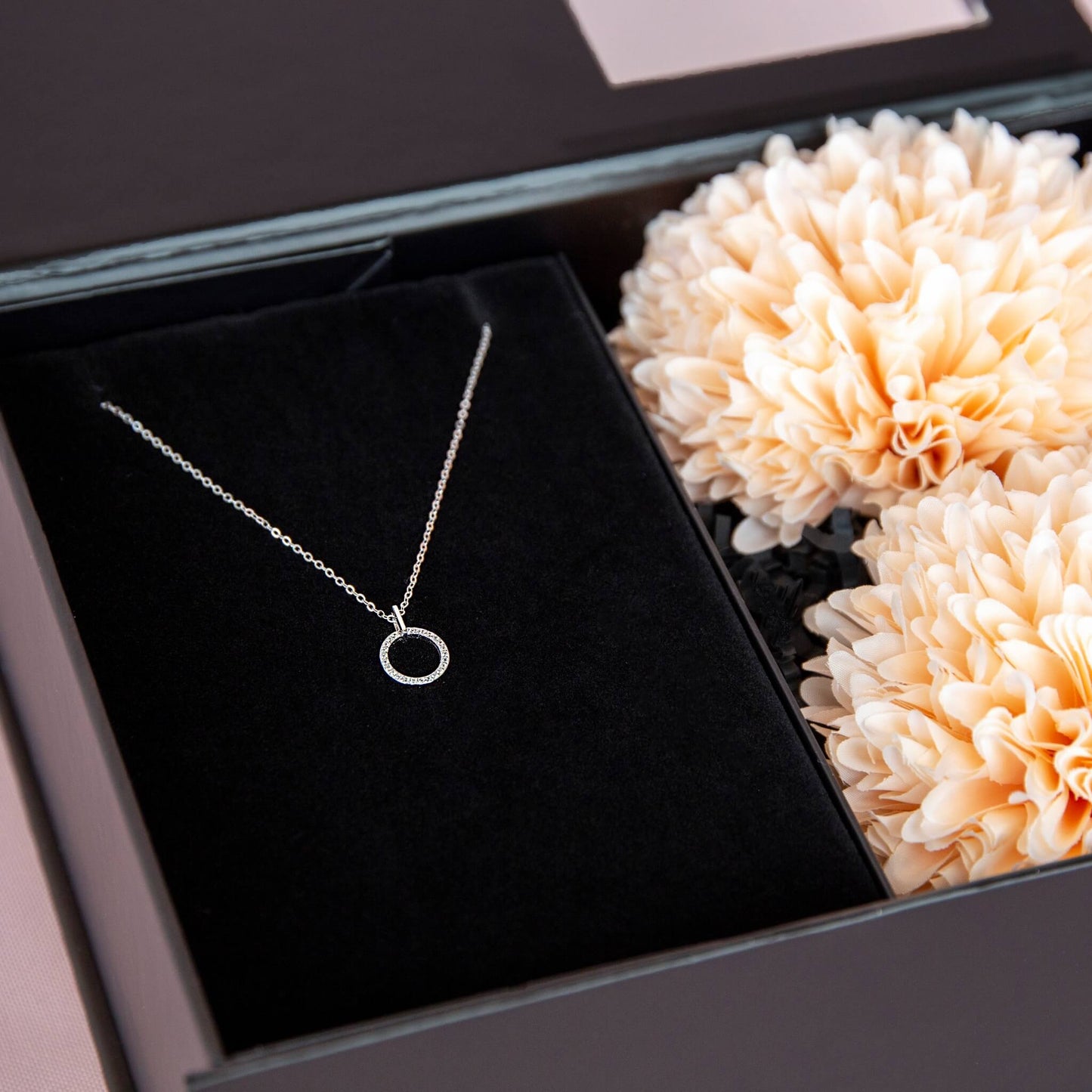 Detlajbilde av rundt smykke i sterling silver med zirkonia stener. Fint plassert i en gaveeske fra Boxit med ferskenfargede blomster.
