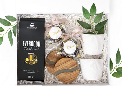 Gave med kaffe, sjokolade, bordbrikker og hvite kopper i en hvit gaveeske.