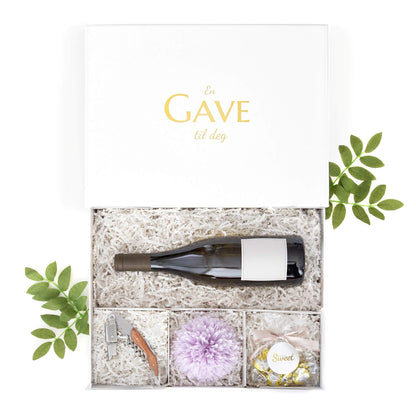 Hvit gaveeske til en vinflaske med lilla blomster, kelneråpner med trehåndtak og sjokolade, en fin innflyttingsgave