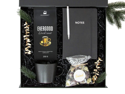 Svart gaveeske med kaffe, kopp, notatbok og sjokolader. Dekorert med juledekor