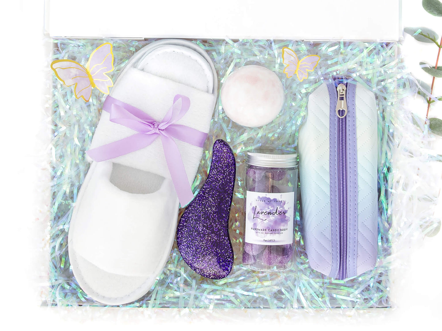 En gaveeske med holografisk fyll som inneholder produkter som slippers, bodyscrub, badebombe, lilla toalettmappe og glitter hårbørste.