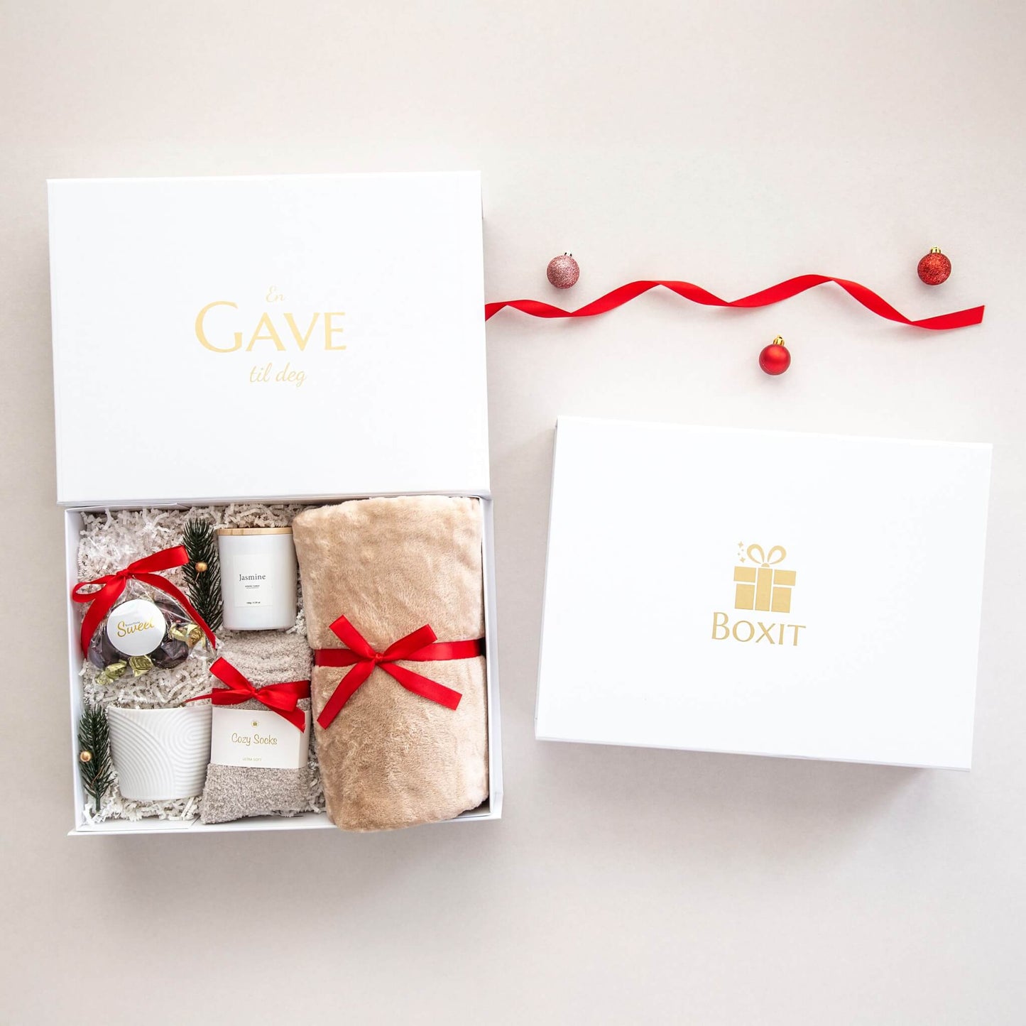 To hvite gaveesker fra Boxit hvor en er lukket og en er åpen og viser et mykt pledd, sokker, duflys, sjokolader og en hvit designkopp. Alt dekorert for jul.