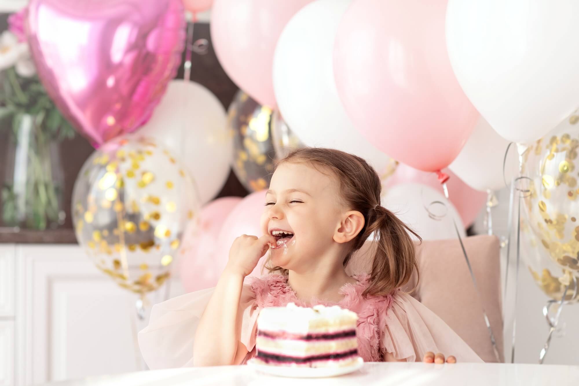 Liten jente spiser bursdagskake omgitt av ballonger på bursdagsfest.