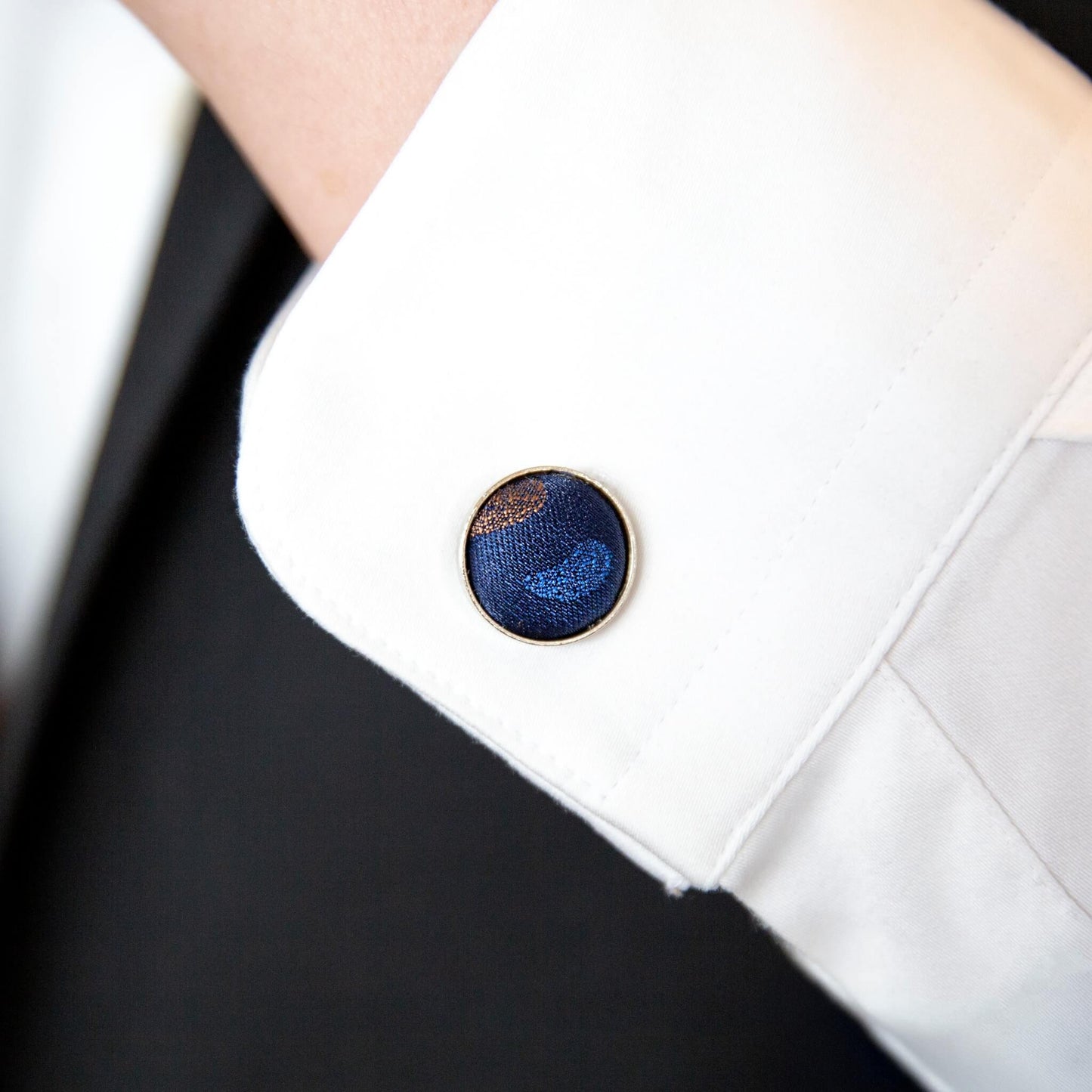 Rund mansjettknapp med blått og kobberfarget stoff brukt på hvit dresskjorte – detalj fra herregavesett.