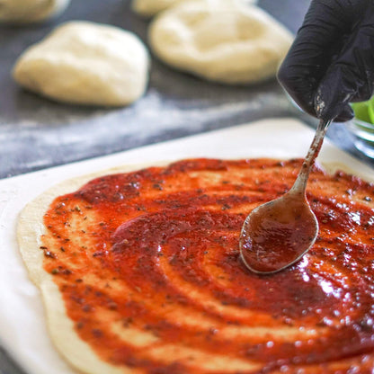 Pizzasaus og krydder som fordeles over en utkjevlet pizzabunn